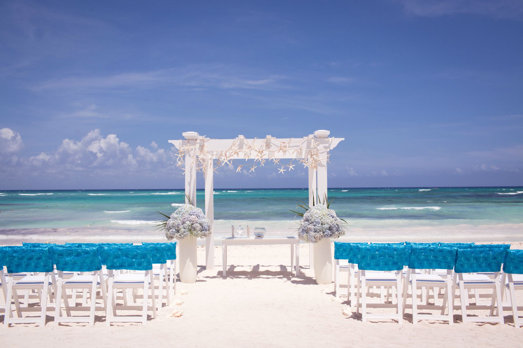 Beach wedding setup with blue-covered white chairs and arch decorated with sand dollars