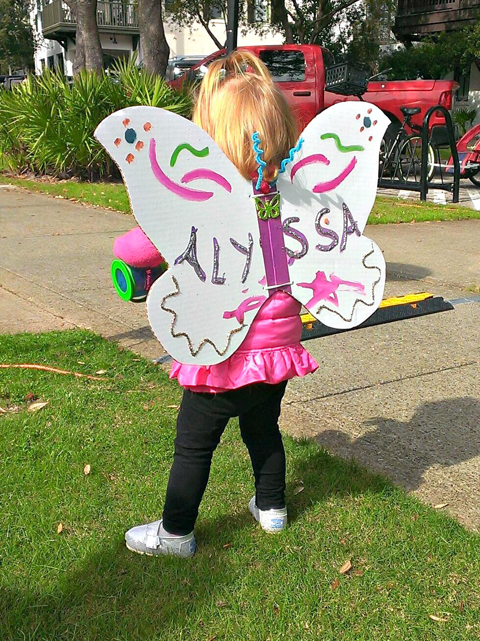 Little girl in pink tunic, black leggings, and posterboard butterfly wings at Fluttery Festival in Rosemary Beach for Highway 30-A arts blog